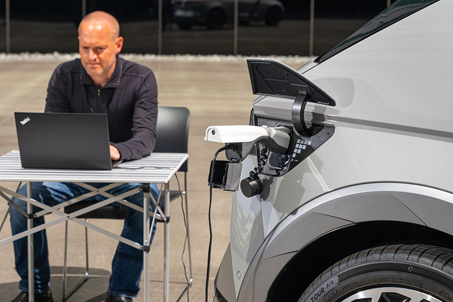 Man working on a laptop powered directly by bidirectional charging from his EV. A person uses a ThinkPad plugged into a white electric SUV via its vehicle-to-load (V2L) port, showcasing how bidirectional-capable EVs can export power to run devices anywhere.