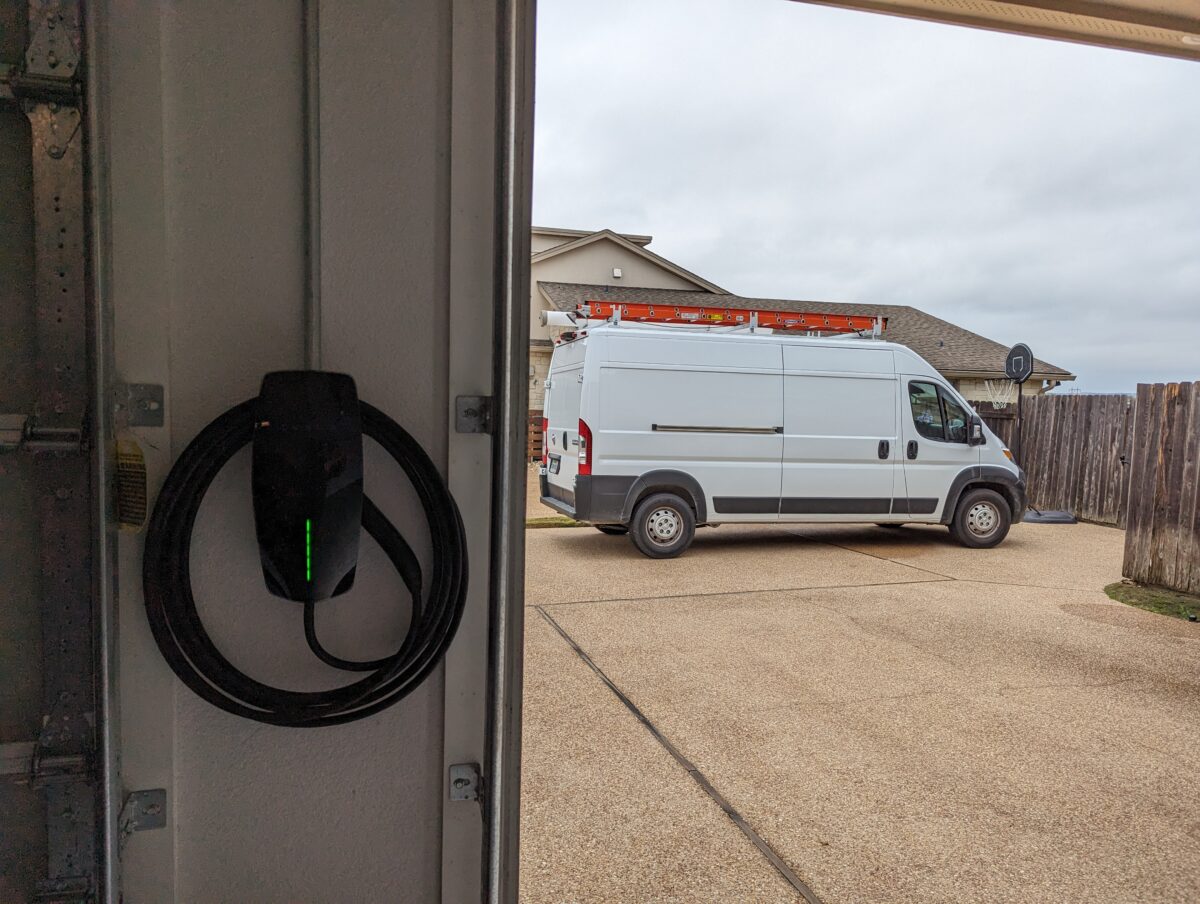 A Tesla Wall Connector EV charger mounted on the interior garage wall next to an open garage door, with its J1772 plug hanging neatly coiled on the holster and a solid green status light indicating it's powered and ready to charge. In the background, a white Ram ProMaster cargo van is parked in the driveway of a suburban Texas home under an overcast sky, with a basketball hoop visible on the right.