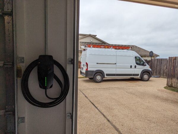 A Tesla Wall Connector EV charger mounted on the interior garage wall next to an open garage door, with its J1772 plug hanging neatly coiled on the holster and a solid green status light indicating it's powered and ready to charge. In the background, a white Ram ProMaster cargo van is parked in the driveway of a suburban Texas home under an overcast sky, with a basketball hoop visible on the right.