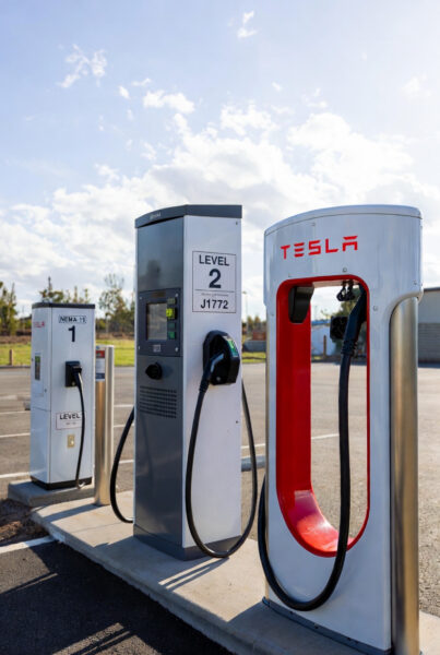 Three electric vehicle charging stations at an outdoor site under a bright sky. In the foreground, a red Tesla Supercharger with its signature curved cable design hangs ready. Behind it are two Level 2 chargers (one labeled LEVEL 2 J1772 and one NEMA 14-50 style) mounted on posts, with black cables plugged in. The chargers are branded with Tesla logos and placed on a concrete pad with grass and trees visible in the background.