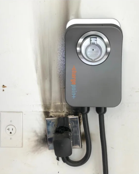 Close-up of a ChargePoint home EV charger plugged into a charred, melted NEMA 14-50 receptacle showing extensive black burn marks, soot, and smoke damage on the surrounding wall from overheating during charging.