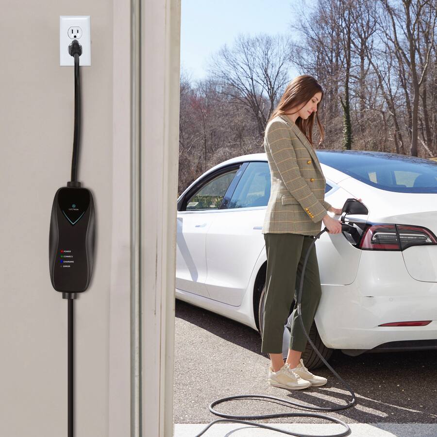 A woman in a plaid blazer and green pants stands next to a white Tesla Model 3 in a residential driveway, plugging in the charging cable. A black Lectron Level 2 EV charger is wall-mounted beside an open garage door, plugged into a standard household outlet (NEMA 14-50 or similar). The charger has indicator lights labeled POWER, CONNECT, CHARGING, and ERROR. Bare winter trees are visible in the sunny background.