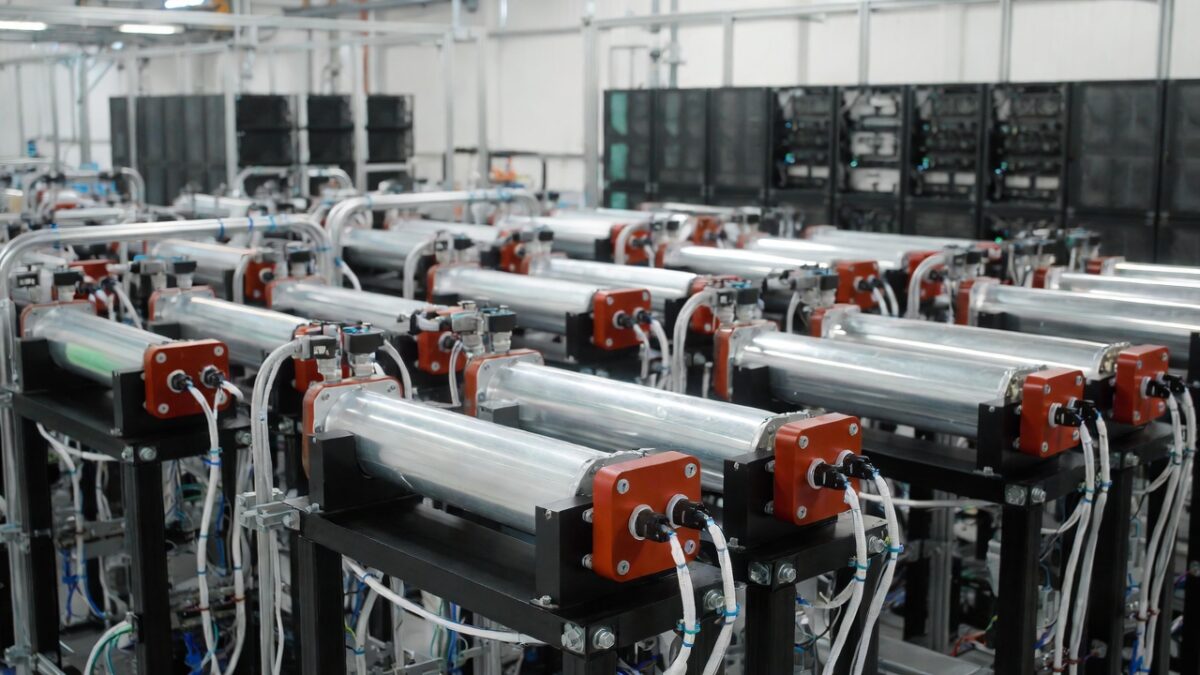 ows of cylindrical sodium-ion battery modules in an advanced U.S. manufacturing facility, featuring silver tubes with red end caps and connected piping, assembled on black metal frames with visible wiring and blue tubing, representing Energy Vault and Peak Energy's partnership for grid-scale energy storage systems.