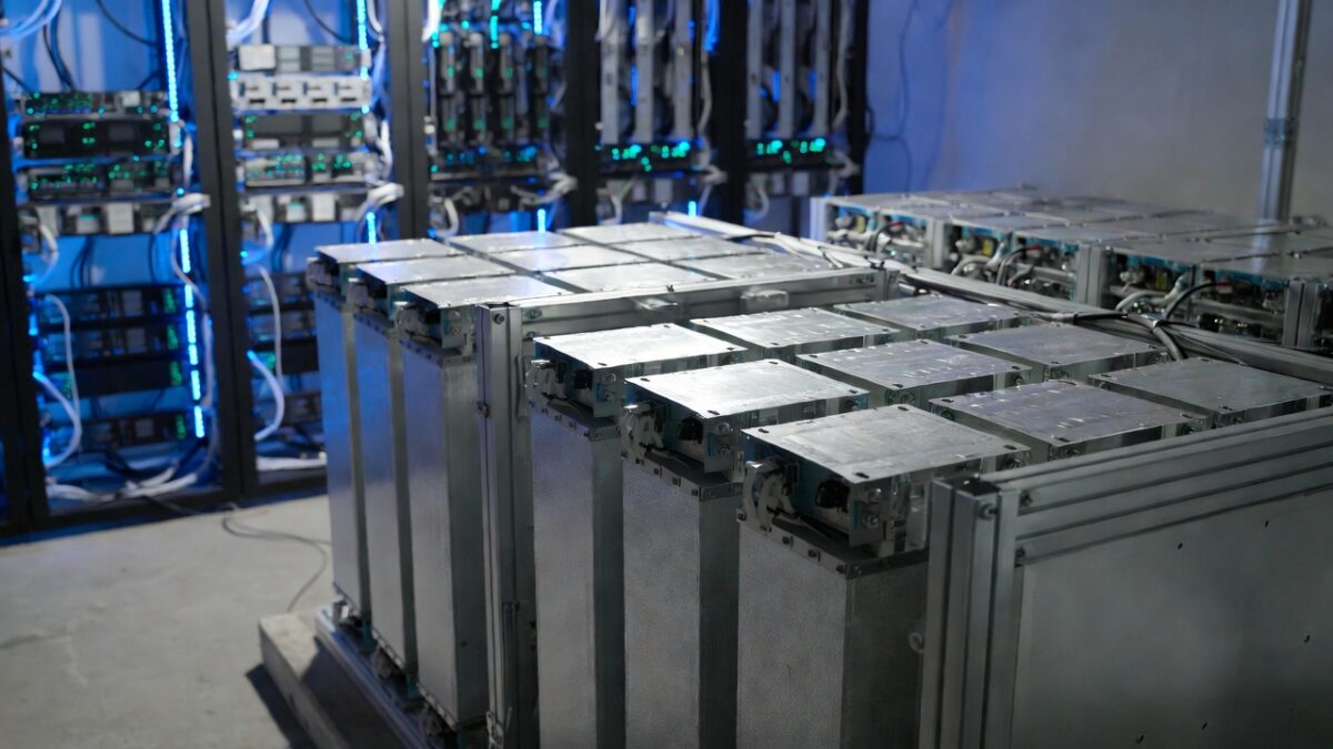 Rows of large rectangular sodium-ion battery modules installed in a high-tech data center environment, featuring metallic silver casings with blue-green LED lighting accents, mounted on racks with visible cooling fans, power connectors, and extensive white cabling, surrounded by server racks glowing with blue lights in the background, illustrating grid-scale or AI data center energy storage deployment.