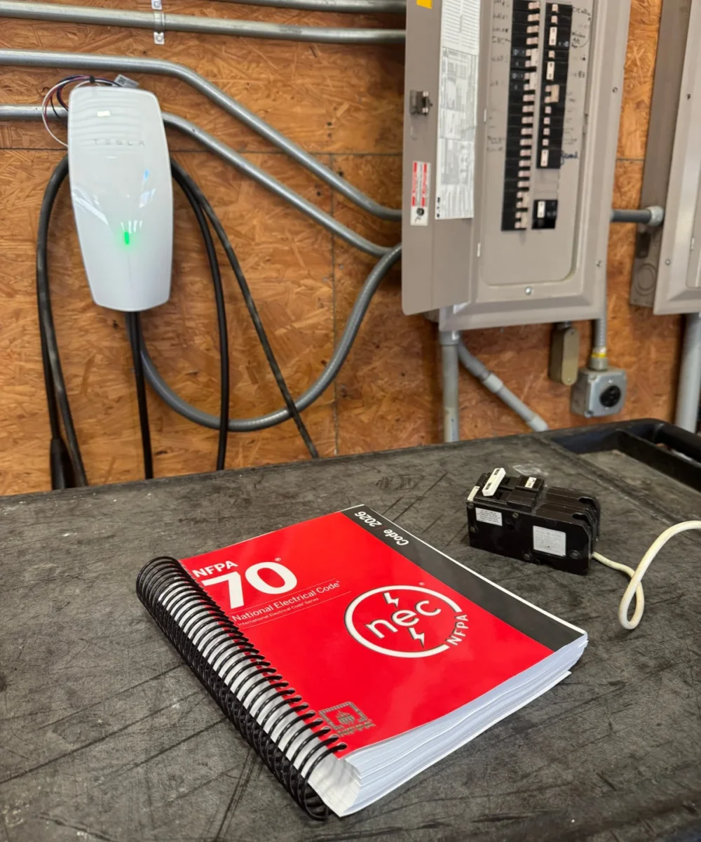 2026 National Electrical Code book and a two-pole GFCI breaker on a workbench in front of a Tesla Wall Connector and electrical panel