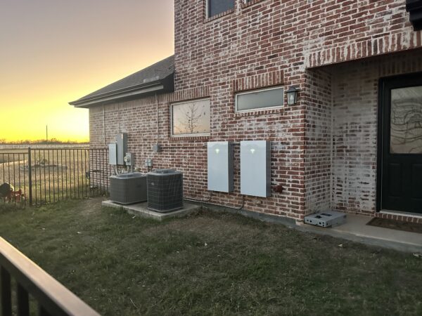 Two Tesla Powerwall 3 batteries on a brick wall at sunset beside the AC units